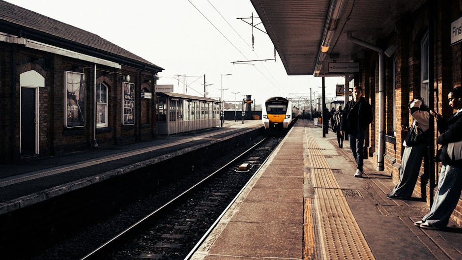 A photograph of a train station platform during daytime shows several passengers waiting for the train, with some standing near the edge while others sit on benches. The platform is paved with concrete and features tactile paving strips along the edge for safety. To the left, a brick station building with posters on the windows is visible, along with a metal awning providing shelter. The train approaching the station is white and yellow, with large windows, and is moving towards the platform. Overhead, electrical wires and signals are strung across the tracks, which are laid with steel rails on wooden sleepers. In the background, additional train tracks and station infrastructure extend into the distance. The scene is well-lit with natural daylight, emphasizing the urban environment typical of house removals and moving logistics, as services like Man With a Van New Malden support streamlined furniture transport and packing during home relocations near the rail network.