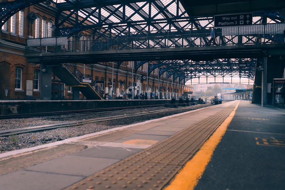 The image depicts a train station platform under an industrial-style metal and glass roof with structural beams overhead. On the platform's left side, there is a brick building with white-framed windows and a staircase leading to an upper level. Several people can be seen standing near the edge of the platform, likely waiting for a train or supervising loading activities. The platform surface consists of paving slabs, with a tactile yellow pathway running along the edge for safety. Adjacent to the platform, train tracks run parallel, and in the background, there are additional station structures and a train car. The scene is well-lit with natural daylight, suggesting a clear day. This setting aligns with house and furniture removals, emphasizing loading and transport processes relevant to relocation services. Man With a Van New Malden provides removals and moving support in this environment, where careful handling of packages, furniture, and boxes is essential for efficient home relocation and furniture transport tasks.