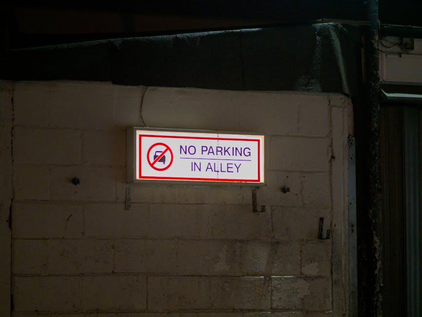 A rectangular illuminated sign mounted on a beige brick wall displays a red circle with a diagonal line crossing out a car icon, indicating no parking allowed. Next to the icon, purple text reads 'NO PARKING IN ALLEY' in capital letters. The sign is positioned above a first-floor window with metal security bars and partially visible ductwork or piping on the right side of the image. The scene is captured at night with minimal ambient lighting, suggesting an urban environment. This signage is relevant for household relocation or furniture transport services, as it indicates parking restrictions in the area, which [COMPANY_NAME], such as Man With a Van New Malden, must consider during home removal and packing processes. The image emphasizes the importance of checking local parking rules before executing a move to avoid fines, aligning with the topic of avoiding fines during house removals in New Malden.