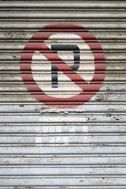 A close-up of a weathered metal roller shutter door painted with a faded 'No Parking' symbol, consisting of a red circle with a diagonal line crossing out a black letter 'P'. In front of the shutter, a moving company representative from Man With a Van New Malden is loading a variety of household items. The items include cardboard boxes of different sizes, some wrapped in plastic and others with packing tape, a wooden furniture piece partially visible, and a vacuum cleaner resting on the pavement. The scene appears to take place outside a residential property, with the items being prepared for home relocation or furniture transport, as part of an organized packing and moving process. The lighting is natural, indicating daytime, and the background suggests a typical urban or suburban street environment with practical logistics involved in house removals.