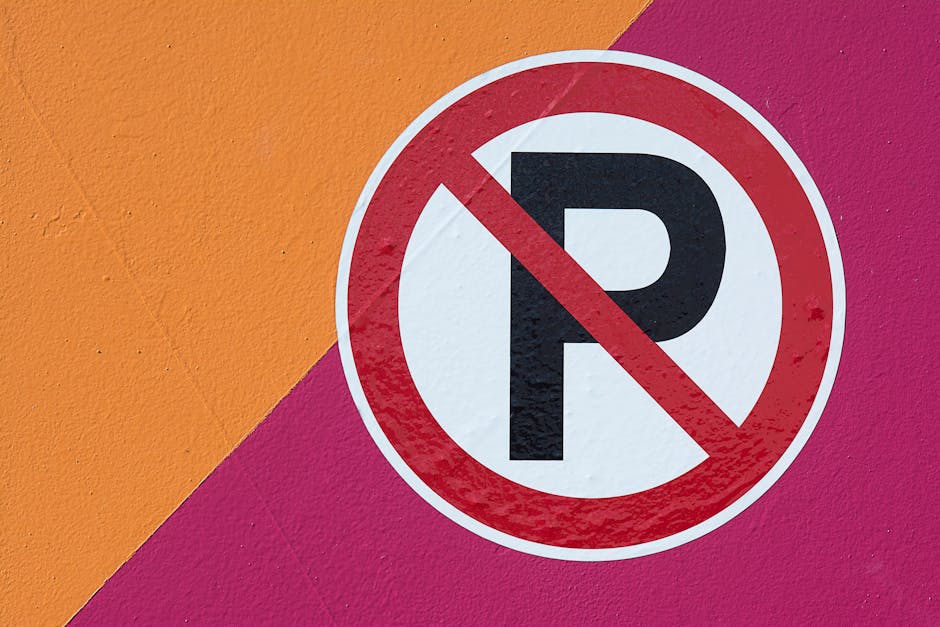 A rectangular illuminated sign mounted on a beige brick wall displays a red circle with a diagonal line crossing out a car icon, indicating no parking allowed. Next to the icon, purple text reads 'NO PARKING IN ALLEY' in capital letters. The sign is positioned above a first-floor window with metal security bars and partially visible ductwork or piping on the right side of the image. The scene is captured at night with minimal ambient lighting, suggesting an urban environment. This signage is relevant for household relocation or furniture transport services, as it indicates parking restrictions in the area, which [COMPANY_NAME], such as Man With a Van New Malden, must consider during home removal and packing processes. The image emphasizes the importance of checking local parking rules before executing a move to avoid fines, aligning with the topic of avoiding fines during house removals in New Malden.