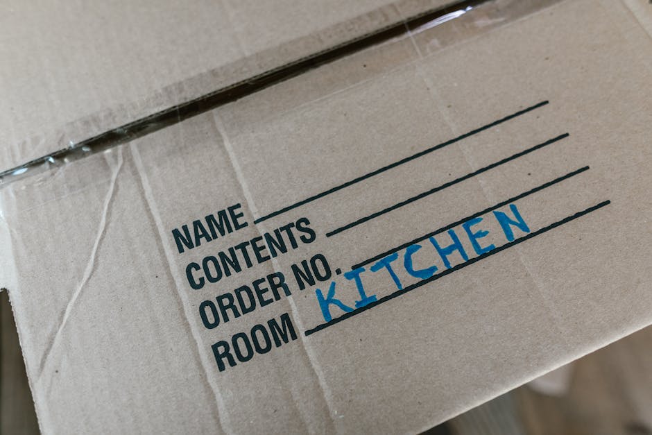 Close-up view of a cardboard moving box with printed labels and handwritten entries, indicating contents for house relocation. The box has tape securing it and features labels for 'NAME,' 'CONTENTS,' 'ORDER NO.,' and 'ROOM,' with the word 'KITCHEN' written in blue marker next to the 'ROOM' label. The box is part of a packing process typically involved in furniture transport and packing and moving services. It rests on a surface inside a property, with part of a doorway visible in the background. The environment appears well-lit, and the focus is on the organized packing materials used by Man With a Van New Malden during the home relocation or house removal process.