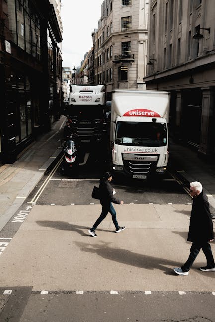 A street scene in an urban area with a narrow road flanked by tall multi-story buildings on both sides. Two delivery trucks, one with the branding 'uniserve' and another with 'Becton Transport,' are parked along the curb, partially blocking the narrow road. A motorcycle is parked on the left side of the street, close to the sidewalk. Two pedestrians are crossing the road on a marked pedestrian crossing; one is a woman dressed in dark clothing carrying a bag, walking from left to right, and the other is an older man in a dark coat walking in the opposite direction. The environment appears overcast with natural daylight illuminating the scene, conveying a typical city moving or delivery operation as part of a home relocation or furniture transport process, with Man With a Van New Malden possibly involved in the logistical activity.
