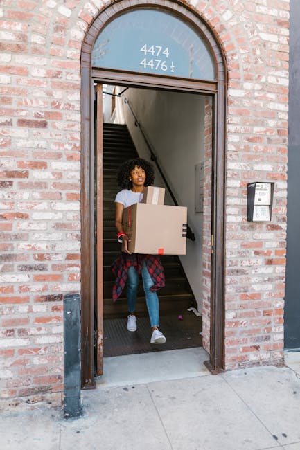 A young woman with curly hair carrying three cardboard boxes, one larger and two smaller, as she emerges from a brick building with an arched doorway. The interior staircase is visible behind her, indicating she is moving items out of the property. She is dressed casually in a light t-shirt, jeans, and sneakers, with a red and black plaid shirt tied around her waist. The building has a black metal post on the pavement near the entrance, and a small black intercom panel is mounted on the brick wall to the right of the doorway. The scene captures the loading process during a home relocation, with natural daylight illuminating the exterior and interior. This image demonstrates the packing and furniture transport aspect of residential removals, likely coordinated by a professional moving service such as Man With a Van New Malden.