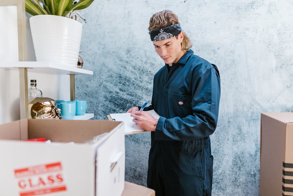 A young male mover wearing a navy blue uniform and a black bandana is inside a property, standing next to a cardboard box filled with various items. He is holding a clipboard and pen, inspecting or noting details about the packing or inventory process as part of a home relocation. To his left, there is a white shelving unit with decorative items such as a large white ceramic pot, a glass bottle, two blue cups, and a gold decorative object. The background features a textured light blue wall, suggesting an organized packing or moving environment. This scene aligns with furniture transport and packing activities involved in professional removals, as provided by Man With a Van New Malden, supporting effective and careful home moving logistics.