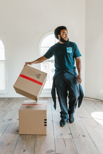 A young woman with curly hair carrying three cardboard boxes, one larger and two smaller, as she emerges from a brick building with an arched doorway. The interior staircase is visible behind her, indicating she is moving items out of the property. She is dressed casually in a light t-shirt, jeans, and sneakers, with a red and black plaid shirt tied around her waist. The building has a black metal post on the pavement near the entrance, and a small black intercom panel is mounted on the brick wall to the right of the doorway. The scene captures the loading process during a home relocation, with natural daylight illuminating the exterior and interior. This image demonstrates the packing and furniture transport aspect of residential removals, likely coordinated by a professional moving service such as Man With a Van New Malden.