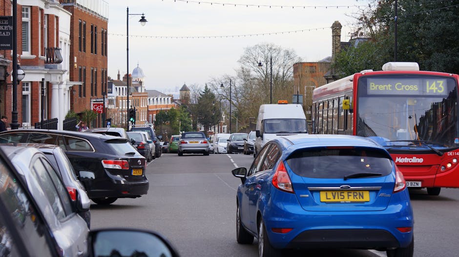 A busy street scene in New Malden featuring several parked vehicles along the roadside, including a black estate car, a silver van, and a blue hatchback with a visible yellow registration plate, parked close to a brick residential building on the left. A red Metroline bus with route number 143 towards Brent Cross is positioned on the right side of the image, partially blocking the view of the pavement. The bus displays the fleet number DE114 on the front. In the background, additional vehicles and trees are visible, with streetlights and overhead utility wires crossing above. The scene is illuminated by natural daylight, with overcast sky and some pedestrians walking or waiting nearby. Within this context, Man With a Van New Malden's home relocation and furniture transport services are often involved in assisting with vehicle permits and loading processes for local house removals, as seen in such urban moving environments.