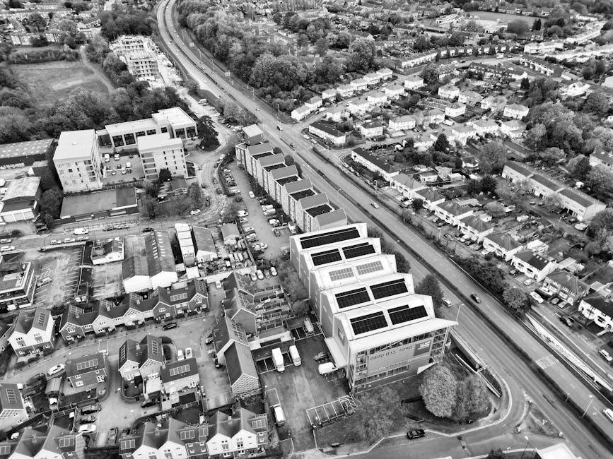 An aerial black-and-white photograph shows a row of modern commercial buildings with flat roofs fitted with multiple solar panels, situated along a busy street in New Malden. In the foreground, a parking area adjacent to the buildings contains several parked cars and small utility vans. Behind the commercial buildings, there is a residential area with rows of terraced houses, some with solar panels on their roofs, surrounded by lush greenery and trees. The street runs parallel to railway tracks visible at the side of the image, with a few cars in motion. The overall scene captures a typical urban environment that may be associated with home and business relocations, as well as furniture transport during home removals or packing and moving processes, with the company Man With a Van New Malden potentially involved in such logistical activities.