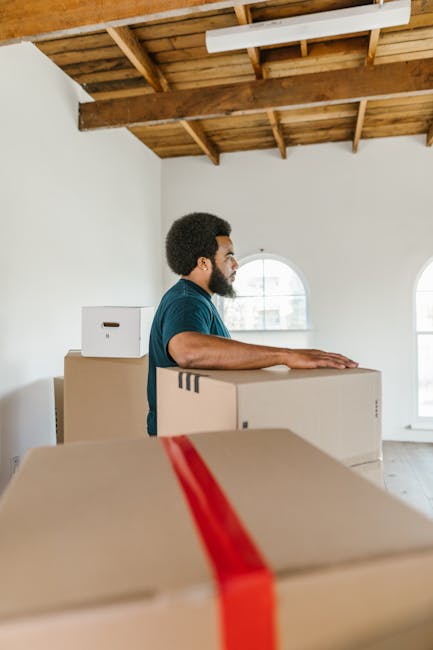 A man with dark curly hair and a beard, dressed in a blue t-shirt, is standing inside a room with white walls, wooden ceiling beams, and large arched windows, during a home relocation. He is positioned behind a stack of cardboard moving boxes, one of which he is resting his hands on, ready for lifting or carrying. The boxes are sealed with packing tape, with some featuring visible cut-out handles for easier handling. Several other boxes are stacked nearby, suggesting a packing or unpacking process related to furniture transport and moving logistics. The room appears well-lit with natural light coming through the windows. The setting indicates a professional removals service, such as Man With a Van New Malden, preparing for the loading process in a home during a house move, with visible elements like the wooden ceiling and minimal furnishings highlighting a typical interior involved in a property relocation, emphasizing careful packing and handling practices.