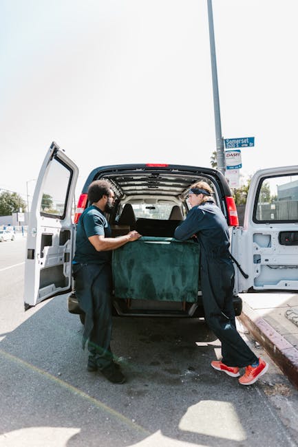 Two removal professionals from Man With a Van New Malden are seen coordinating during a home relocation process, standing beside an open rear door of a white van parked on a street. One worker, wearing a blue shirt, is gripping the edge of a large, green upholstered piece of furniture, while the other, dressed in black with a cap and glasses, is stabilising the item as it is being loaded or unloaded. Inside the van, visible are several cardboard boxes and wrapped furniture pieces, indicating the packing and moving activities. The van's interior has a high roof to accommodate large items, and both workers are using their hands to carefully handle the furniture, ensuring a safe transport. The surrounding environment includes a lamppost with signage, a grey pavement curb, and a partly cloudy sky, reflecting a typical urban moving scene in New Malden. This image captures the logistics of furniture transport as part of a professional removals service, supporting the concept of efficient packing and loading for house relocations.