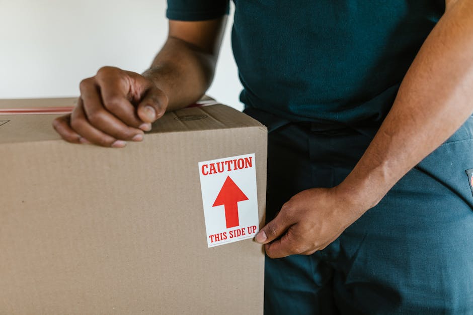 A man with dark curly hair and a beard, dressed in a blue t-shirt, is standing inside a room with white walls, wooden ceiling beams, and large arched windows, during a home relocation. He is positioned behind a stack of cardboard moving boxes, one of which he is resting his hands on, ready for lifting or carrying. The boxes are sealed with packing tape, with some featuring visible cut-out handles for easier handling. Several other boxes are stacked nearby, suggesting a packing or unpacking process related to furniture transport and moving logistics. The room appears well-lit with natural light coming through the windows. The setting indicates a professional removals service, such as Man With a Van New Malden, preparing for the loading process in a home during a house move, with visible elements like the wooden ceiling and minimal furnishings highlighting a typical interior involved in a property relocation, emphasizing careful packing and handling practices.