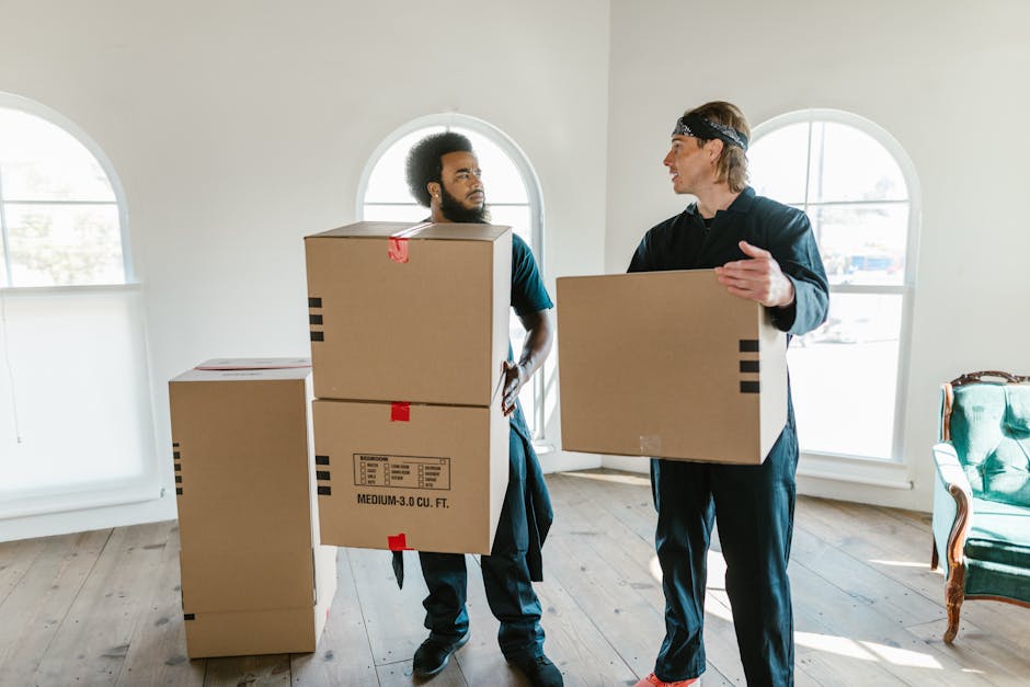 Two movers from Man With a Van New Malden engaged in a home relocation process indoors, each carrying a medium-sized cardboard box. The room has natural daylight streaming through two arched windows, illuminating a wooden floor and light-colored walls. One mover, with curly hair and a beard, is holding two stacked cardboard boxes, secured with red tape, and standing next to additional packed boxes placed on the floor. The other mover, with straight hair and wearing a headband, is holding a single cardboard box, engaging in a discussion, possibly about the loading process. The scene captures the packing and furniture transport stage of a professional removals service, with minimal furnishings visible, including a vintage-style green armchair on the right. The environment suggests an ongoing house move, with boxes ready for transportation, and the background indicates a spacious, well-lit interior typical of a residential property in New Malden.