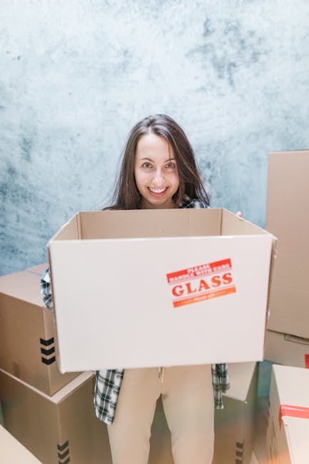 A woman with long brown hair smiling while holding an open cardboard box labeled 'FRAGILE GLASS' in red text, standing in a room with a textured light blue wall. Surrounding her are several other closed cardboard boxes of various sizes, some with black tape securing the lids. The boxes are placed on a flat surface, and she is dressed in casual clothing including a checked shirt and beige pants. The scene depicts the packing phase of home relocation or moving process, with items prepared for transport. This image relates to house removals and furniture transport, highlighting packing and loading activities performed by [COMPANY_NAME] as part of a comprehensive removals service, suitable for last-minute or same-day moves in New Malden.