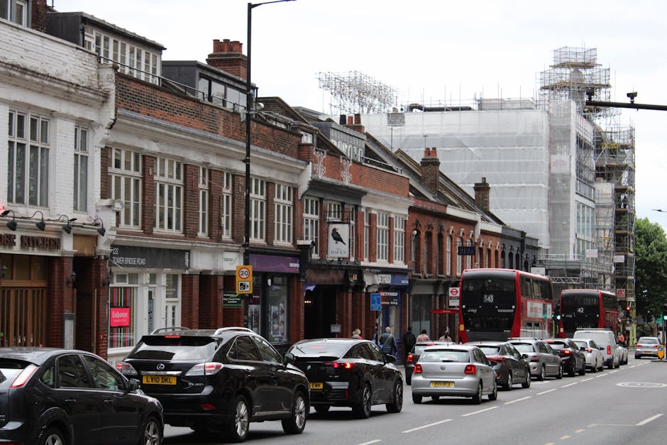 A street scene on Kingston Road, New Malden, showing a line of multi-storey brick and white facaded buildings, some with storefronts such as a cake shop and a bookstore. In front of the buildings, a single lane of parked cars and moving vehicles, including a black Mercedes SUV, several sedans, and two red double-decker buses numbered 343 and 42, are visible. Pedestrians are walking along the sidewalk, some near shop entrances. To the right, a building under construction is covered with scaffolding and white protective sheeting, indicating ongoing building work. The sky above is overcast, and the street appears busy with both vehicles and pedestrians, reflecting typical urban activity associated with home relocation or moving services like those offered by Man With a Van New Malden. The image captures the process of furniture transport or moving logistics in an active city environment.