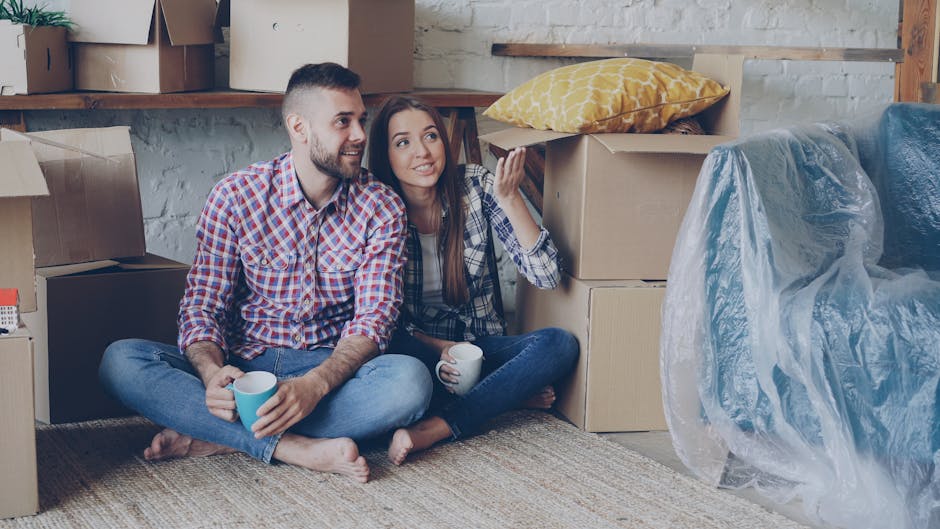 A young couple sits on the floor of a room filled with moving boxes during a home relocation process. The man, wearing a red and blue checkered shirt and jeans, holds a light blue mug, while the woman, dressed in a similar checkered shirt and dark jeans, also has a mug in her hand and appears to be talking and gesturing with her other hand. Around them are several cardboard boxes, some open and some closed, with one box containing a yellow patterned pillow. A large piece of furniture covered in semi-transparent plastic wrap is visible to the right, indicating protection for transport. The background includes a wooden shelf above the boxes, and the setting appears to be an indoor space on Kingston Road, New Malden, during the process of packing and preparing for an upcoming move. This scene is managed by Man With a Van New Malden, specialists in removals and furniture transport, demonstrating packing, loading, and interior organization in a typical house move situation.