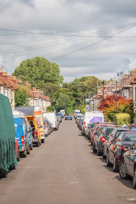 A residential street during daytime with parallel-parked vehicles on both sides, including cars and small vans, some with covers or protective sheeting in various colors. The street is lined with terraced houses featuring brick facades, some with front gardens or small trees, one with vibrant red and green foliage. In the background, there are larger trees with lush green foliage and a cloudy sky overhead. Overhead power lines run across the street. This scene depicts a typical urban area suitable for home relocation or furniture transport, with some vehicles possibly used by [COMPANY_NAME] for moving services. The calm environment and organized parking make it ideal for detailed loading and unloading activities during a house removal, as part of a professional packing and moving process documented on the New Malden Broadway Moving Guide: Narrow Streets Tips, available at [PAGE_URL].