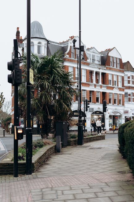 Photograph of a busy street scene outside a classic Victorian-style building with white and red brickwork, large windows, and a rounded turret roof in New Malden. The pavement in the foreground features a pedestrian crossing signal and black lampposts with traffic lights attached. There are some palm-like trees and greenery along the sidewalk, with a few pedestrians visible in the background. The image captures a typical street scene during daylight, with overcast skies, illustrating an urban environment suitable for house removals and home relocation logistics, which Man With a Van New Malden can assist with through furniture transport and packing and moving services.