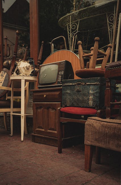 A collection of various used furniture items, including a small white wooden side table, an old television set with a curved screen, a wooden cabinet, and several wooden dining chairs with fabric upholstered seats, all arranged closely together inside a building or storage area. Some chairs are stacked or leaning against each other. The furniture is in different conditions, with visible signs of wear and tear, and the setting is illuminated by natural light from a nearby window or door, suggesting it is part of a house clearance or furniture disposal process. This arrangement is typical of furniture ready for transport or disposal as part of home relocation or clearance services offered by Man With a Van New Malden.