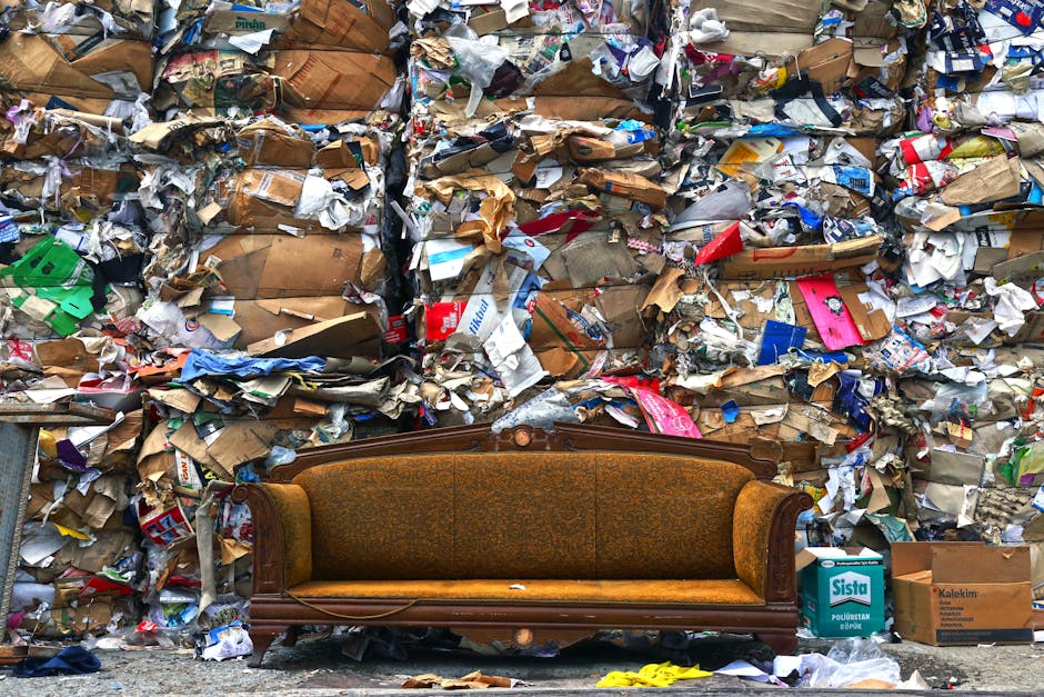 A vintage upholstered wooden sofa with ornate carvings on the armrests and backrest is positioned outdoors on a sidewalk in front of a large, chaotic pile of mixed recycling and waste materials, including crumpled cardboard boxes, paper, plastic packaging, and other refuse. The background features a dense, untidy wall of discarded materials, emphasizing improper disposal or waste accumulation. The scene highlights the importance of proper waste management and furniture disposal regulations relevant to house removals and furniture transport, with the sofa awaiting collection or recycling as part of a home relocation process. The lighting is natural, casting minimal shadows, and the environment suggests an urban area where proper disposal laws are crucial for legal compliance and environmental responsibility, especially in the context of furniture disposal laws for Kingston and New Malden. Man With a Van New Malden provides professional removals services to assist with safe and lawful furniture disposal and home moves.