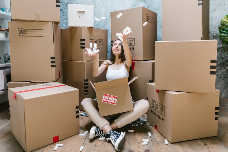 A woman sitting cross-legged on a wooden floor inside a home during packing and moving, surrounded by stacked cardboard boxes of various sizes, some labeled with fragile or contents information. She i