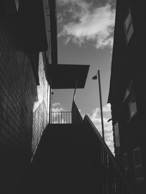 A black and white photograph showing the exterior staircase of a residential building in New Malden during daylight hours. The staircase, constructed of metal with a railing, ascends in a narrow outdoor space between two multi-storey brick buildings. To the left, the building's brick wall is visible, while on the right, another building features windows and a downspout. At the top of the staircase, there is a small landing with a metal railing, leading to an upper doorway or balcony. A streetlamp is mounted on a pole along the side of the building, and the sky above shows partly cloudy conditions with patches of sunlight filtering through. This scene appears relevant to home relocation or furniture transport, consistent with the services provided by Man With a Van New Malden, within a context of loading or preparing for moving or packing activities.