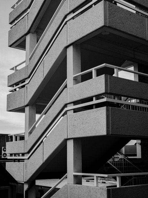 A black and white photograph of a multi-storey residential building's exterior staircase structure, with concrete steps and metal railings. The staircase is arranged in a zigzag pattern, connecting different floors and leading to external walkways with safety railings. The building's exterior is finished with textured concrete panels, and the staircase is supported by vertical concrete pillars. The scene is captured during daylight, with the sky visible in the background. In the context of home relocation and moving services by Man With a Van New Malden, this image illustrates the external access points used for furniture transport and the logistical considerations of loading items from a building's exterior staircase during a house removal or heavy move.