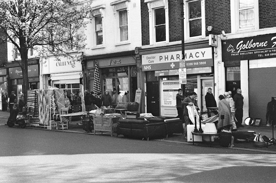 A black and white photograph depicts a busy street scene outside a row of shops, including a pharmacy, with a group of people gathered around furniture and household items displayed on the pavement. Various items such as sofas, boxed goods, wooden chairs, and clothing racks are arranged along the sidewalk, some covered with protective blankets and plastic wrapping. Several individuals are seen carrying or lifting furniture, while others stand nearby observing or assisting with loading. A small van or moving vehicle is partially visible behind the group, suggesting active furniture transport and home relocation processes. The environment appears to be during daytime, with natural light illuminating the scene. Trees line the street, and the storefronts include signage for retail and service businesses. This scene highlights the logistics of packing, moving, or clearing household items as part of a household removal or furniture transport service, consistent with the services offered by Man With a Van New Malden.
