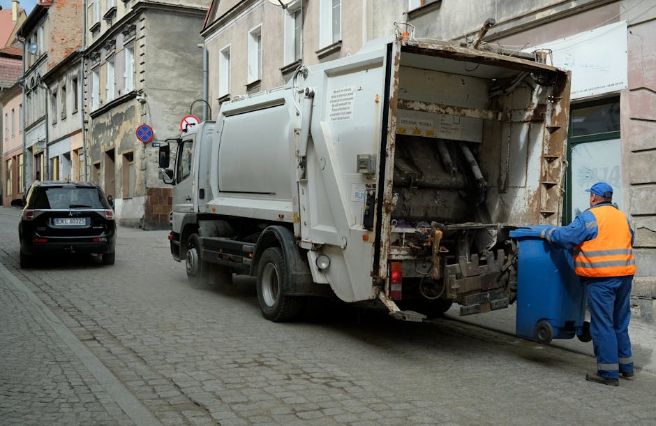 A street scene showing a waste collection vehicle operated by a worker in a blue uniform and orange high-visibility vest, who is loading a blue wheeled rubbish bin into the rear compartment of a white garbage truck. The truck is parked on a cobblestone road beside a building with multiple stories, with several windows and a storefront on the ground level. The rear of the truck is open, revealing mechanical lifting equipment, and dust or debris is visible around the loading area. A black car is parked further along the street, and nearby traffic signs are displayed on lampposts. The scene takes place during daylight hours, with natural lighting illuminating the urban environment. This image illustrates waste collection related to home or commercial property management, which can be connected to house removals or relocation services provided by [COMPANY_NAME], supporting proper disposal during the moving process.